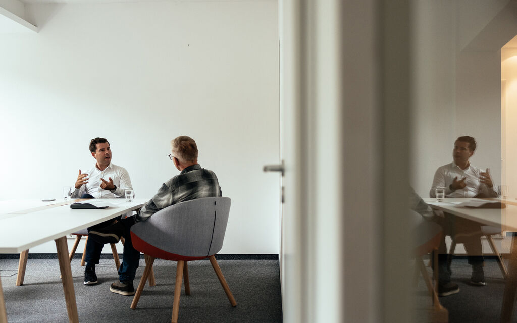 Felix Haas und Stefan Ruzas sitzen in einem Meetingraum auf grauen Stühlen an einem langen weißen Tisch