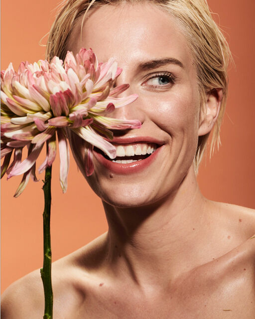 Nahaufnahme einer natürlich geschminkten und lachenden Frau mit Blüte vor dem rechten Auge.