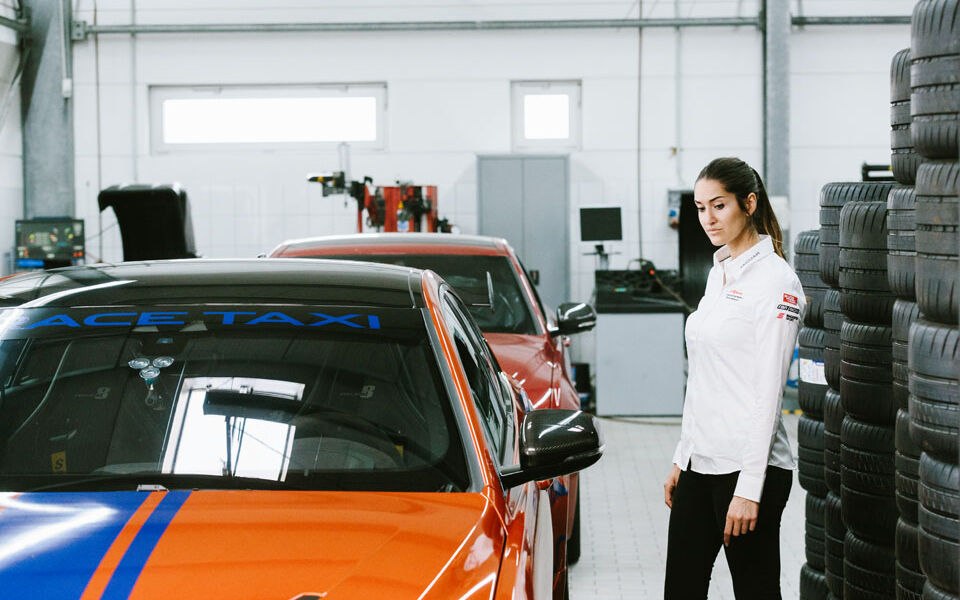 Racing driver Célia Martin stands next to an orange and blue Jaguar racing car in a garage