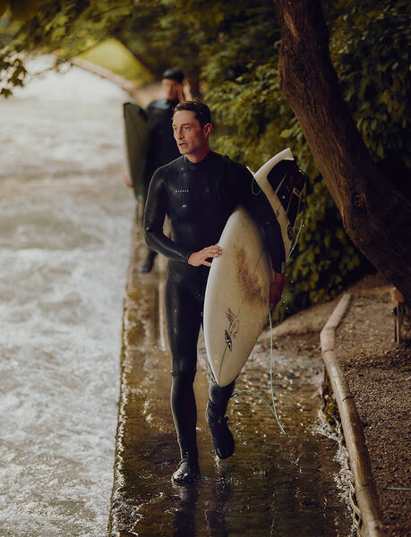 Sebastian Kuhn steht neben dem Eisbach mit Neoprenanzug und Surfbrett in der Hand