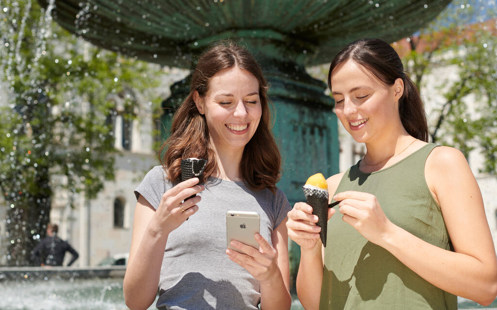 Two young women standing in front of a fountain, eating ice cream and looking at a smartphone.