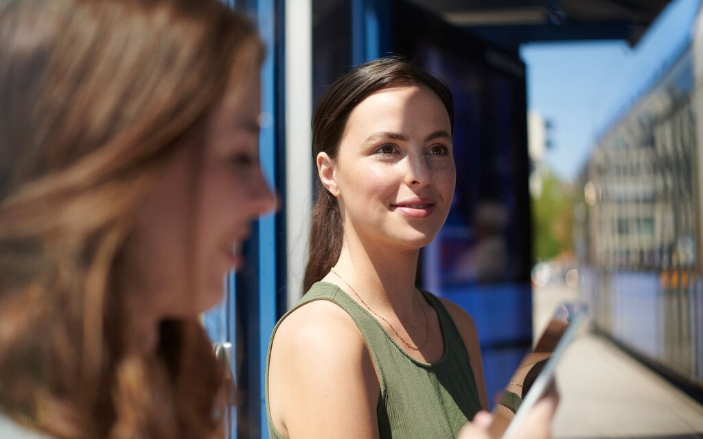 Young woman sitting at a tram station, in the foreground a woman looking at her smartphone