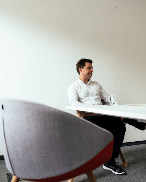 Ein grauer runder Stuhl, im Hintergrund sitzt Felix Haas am Tisch