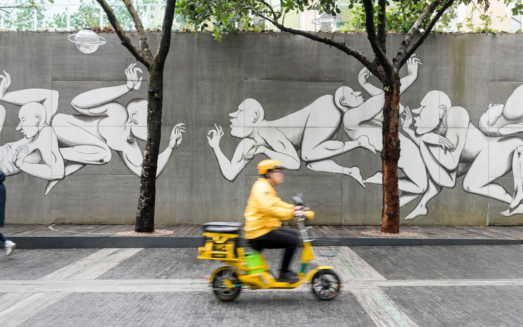 Gelb gekleideter Rollerfahrer fährt in Shenzhen an mit Graffitikunst besprühter Betonwand vorbei