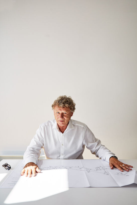 Hermann Tilke sitzt mit weißem Hemd an seinem Schreibtisch mit ausgebreiteten Streckenplan vor sich