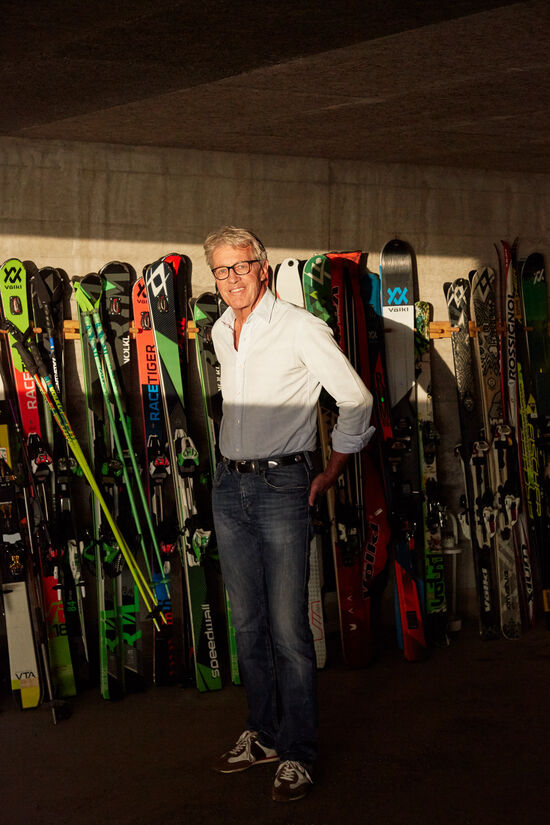 Bernhard Russi stands in front of a wall with several skis and ski poles.