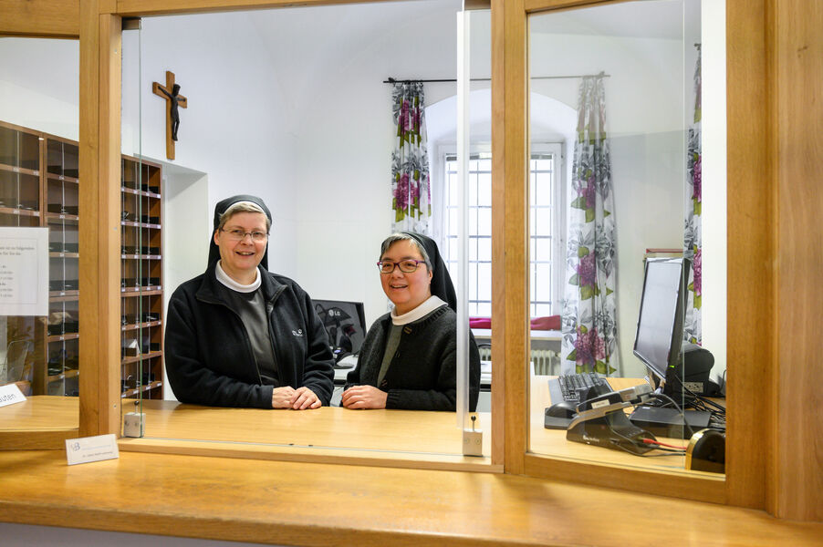 Two nuns stand at the reception of a monastery behind a glass pane