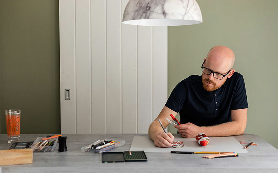 Howard Lee sits at the table and draws chocolate-nicolas, around him pencils and other drawing utensils