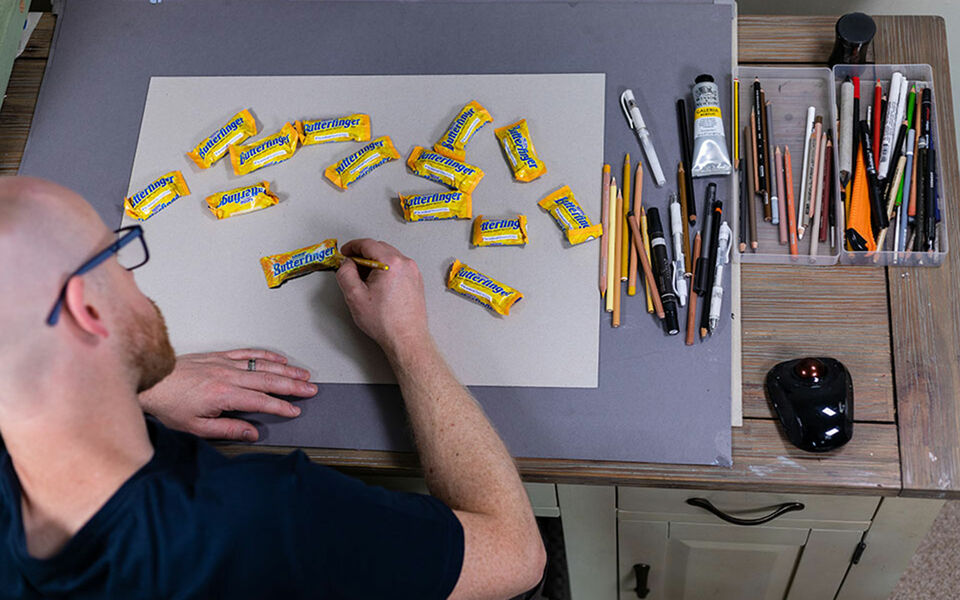 Howard Lee zeichnet gelbe Butterfinger-Schokoriegel ab, neben ihm Stifte und weitere Zeichenutensilien