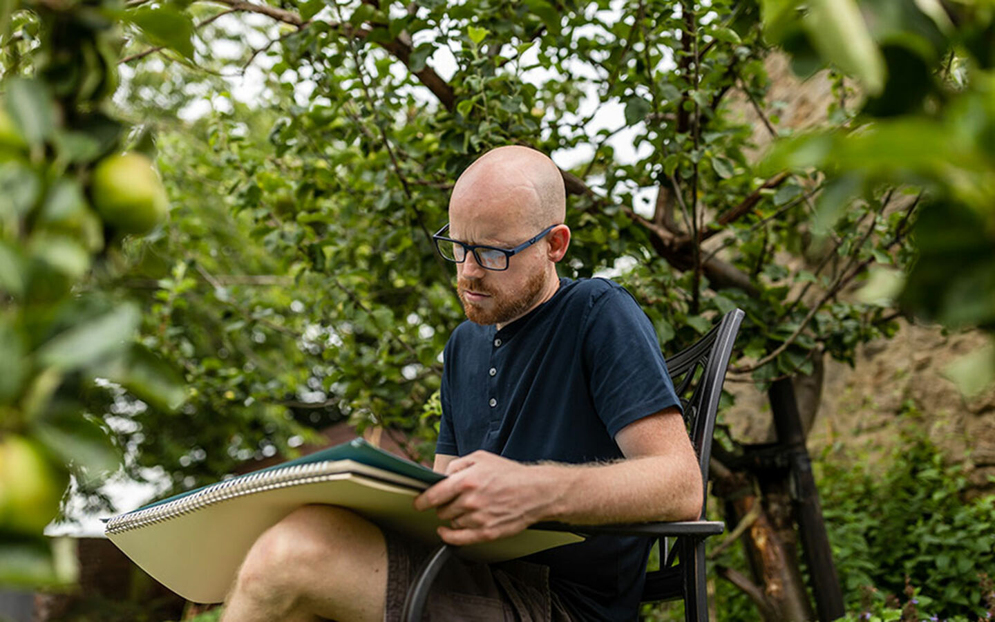 Howard Lee sitzt im Garten auf einer Bank unter Apfelbaum und zeichnet auf Skizzenblock