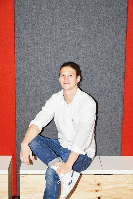 SWM-Werkstudent Simon B sitzt vor einer rot-grauen Wand auf einer Holzbank