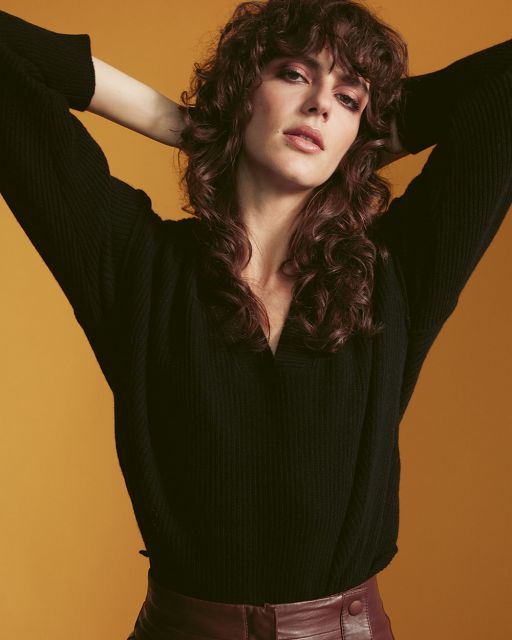 Junge Frau mit braunen Locken mit den Armen hinter dem Kopf trägt einen schwarzen Pullover und einen weinroten Lederrock