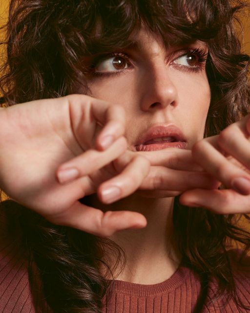 Young woman with brown curls, Smokey Eyes in rust red holds her fingers crossed in front of her mouth