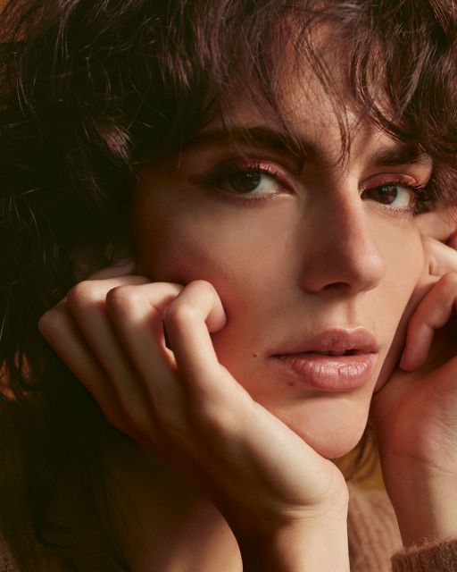 Young woman with brown curls, Smokey Eyes in rust red holds her fingers on her chin