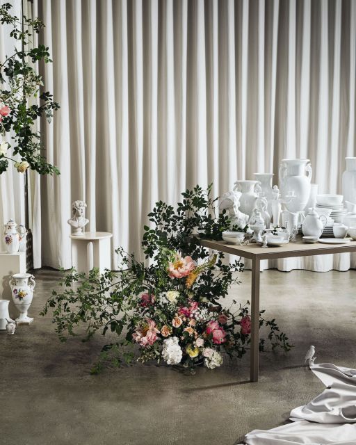 Blumenarrangement vor Tisch mit weißem Geschirr und cremefarbenem Vorhang