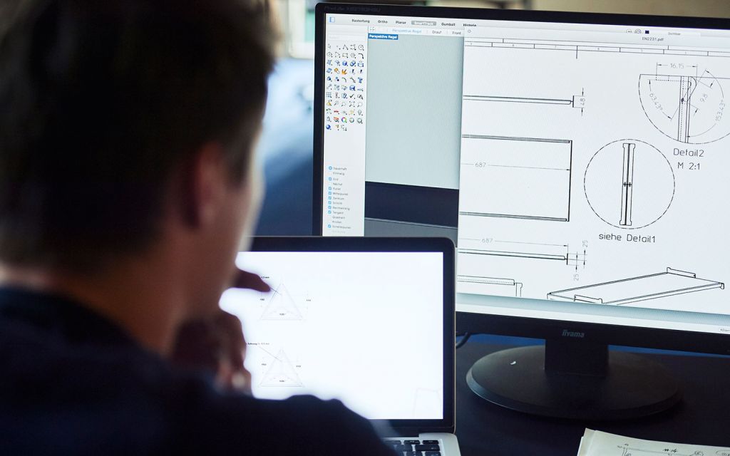 Person sitzt an PC mit Konstruktionsskizzen