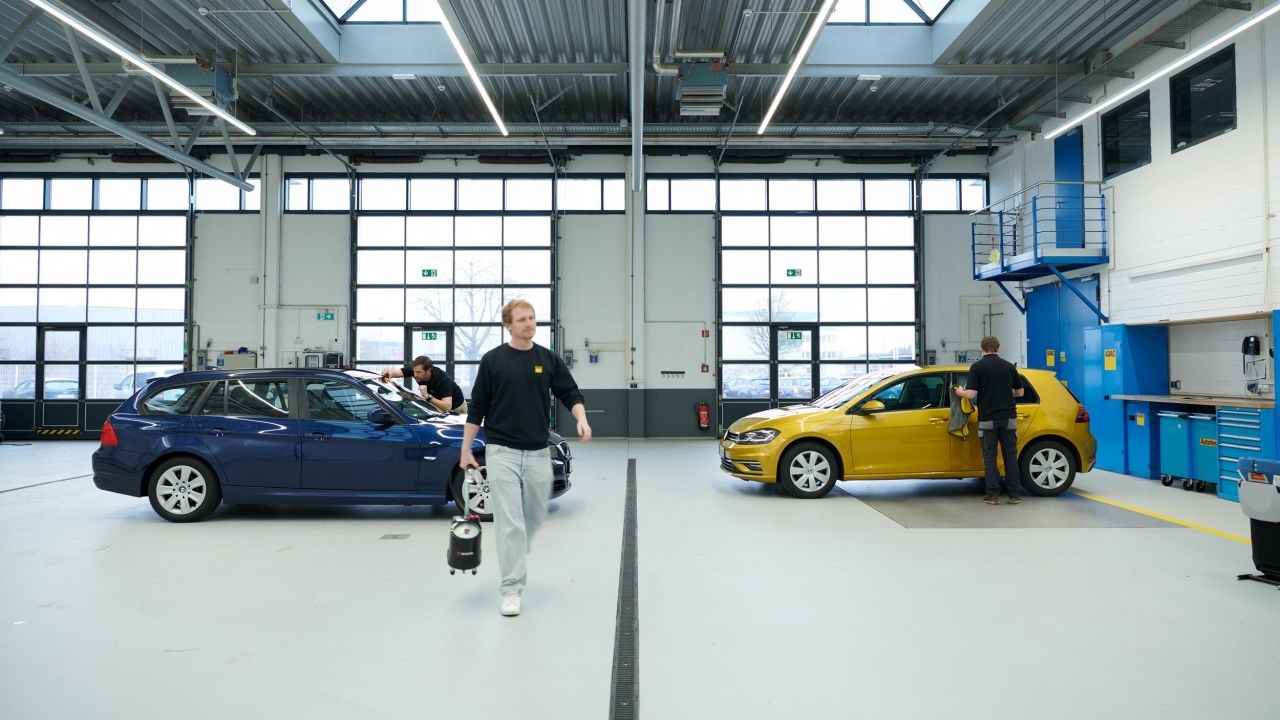 Drei Mechaniker arbeiten in einer hellen Werkstatthalle an einem blauen und einem gelben Kompaktwagen