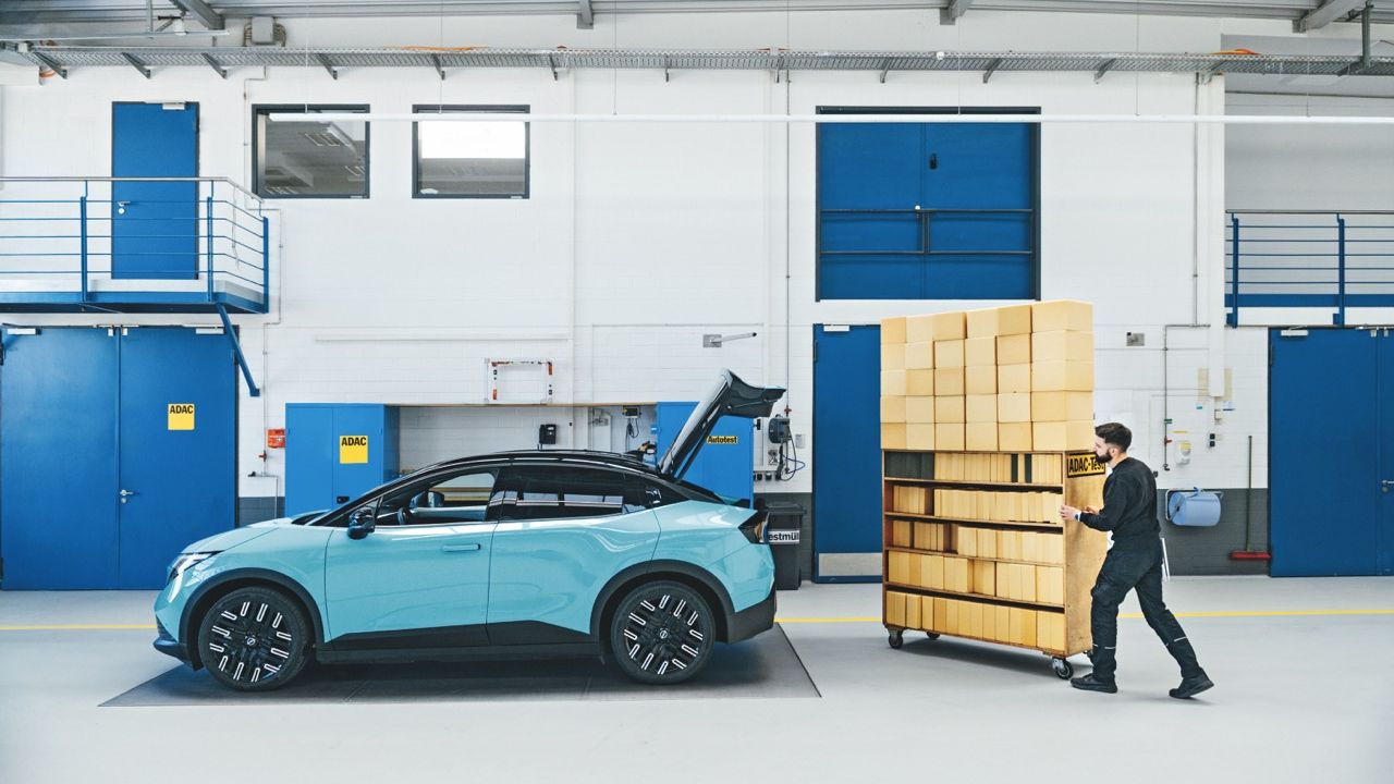 Ein Mann schiebt in einer Fahrzeughalle ein mobiles Regal voller Kisten hinter das geöffnete Heck eines hellblauen Autos.