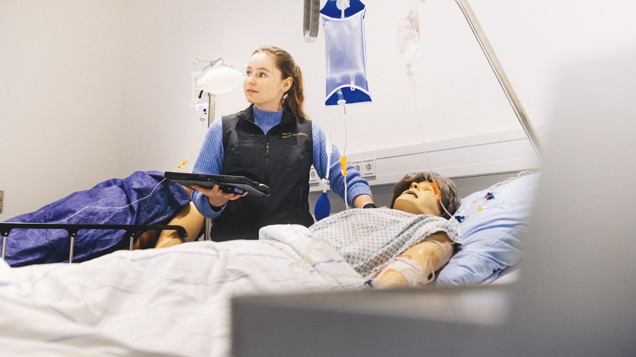 Eine Frau mit einem Tablet steht in einem medizinischen Schulungsraum neben einer Übungspuppe im Krankenbett.