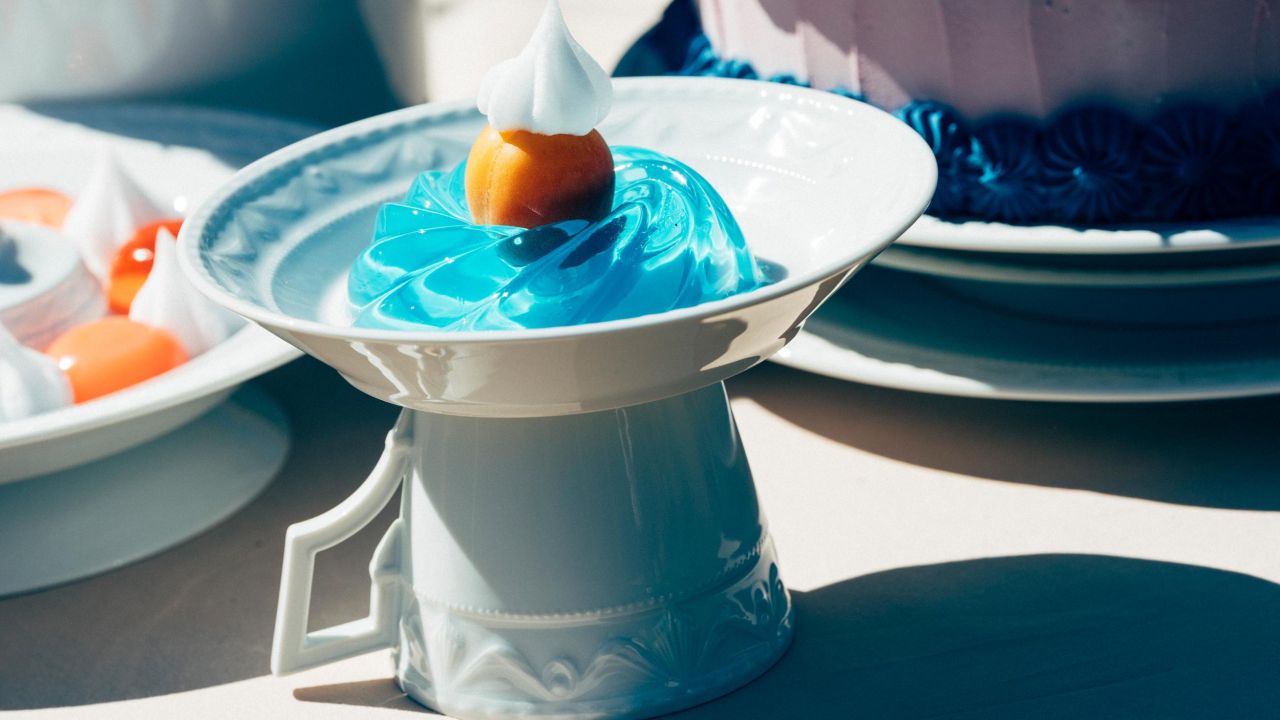 Nahaufnahme eines Tellers mit einem blauen Wackelpudding und einer Frucht, der auf einer umgedrehten Tasse steht. Nahaufnahme eines Tellers mit einem blauen Wackelpudding und einer Frucht, der auf einer umgedrehten Tasse steht.