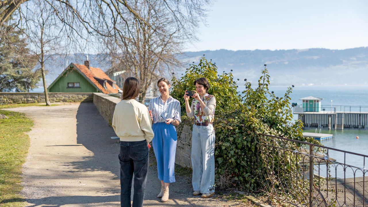 Three women are conversing on a lakeside path with a view of a lake and mountains in the background Three women are conversing on a lakeside path with a view of a lake and mountains in the background