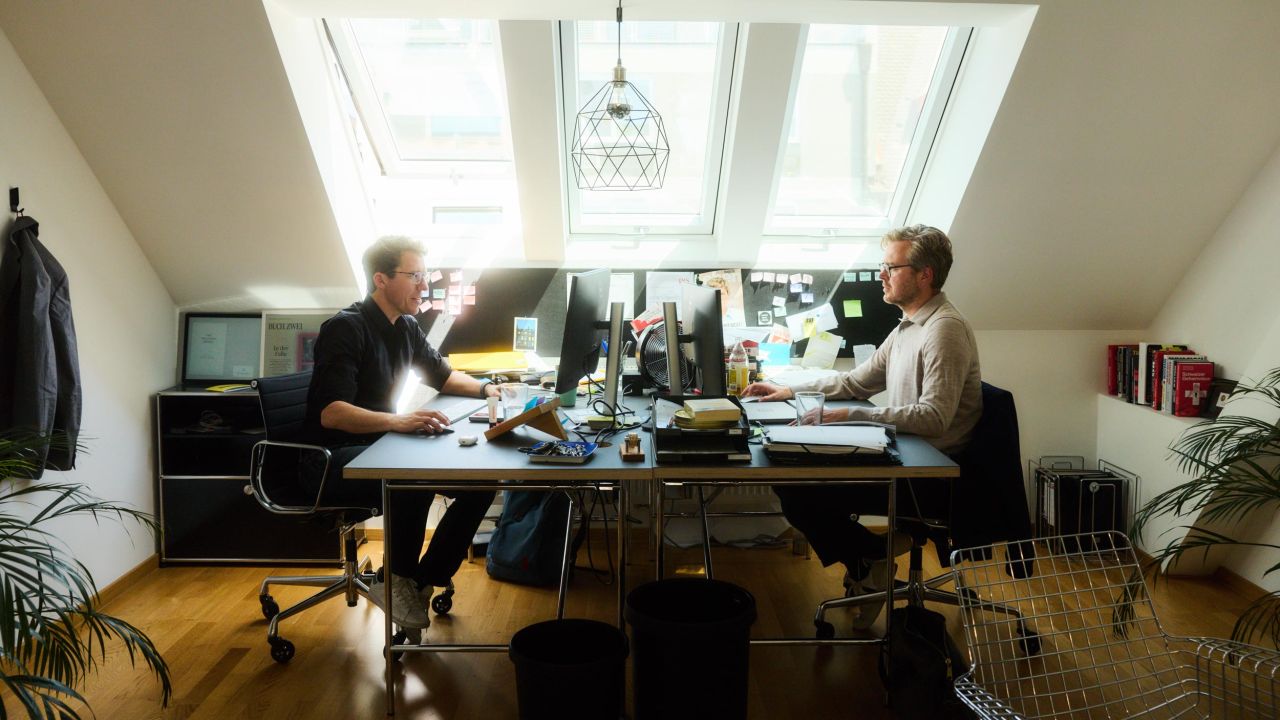 Two men are sitting across from each other at a desk with several monitors in an office. Two men are sitting across from each other at a desk with several monitors in an office.