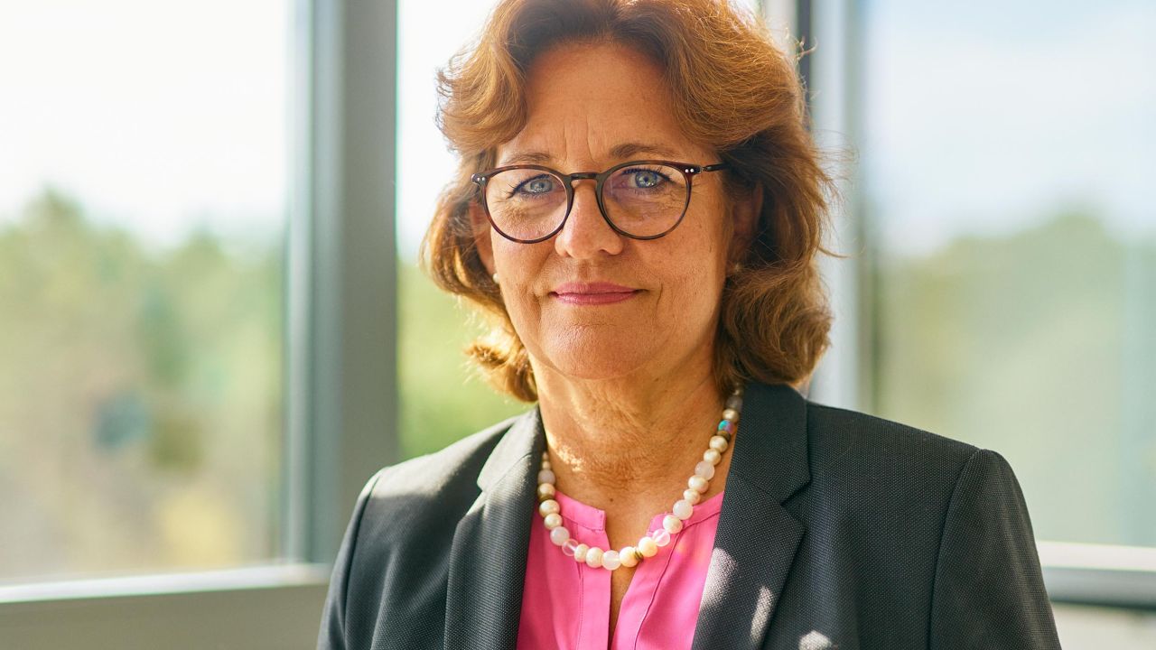 Nahaufnahme einer lächelnden Frau in Businesskleidung mit Brille und Perlenkette vor einem großen Fenster Nahaufnahme einer lächelnden Frau in Businesskleidung mit Brille und Perlenkette vor einem großen Fenster