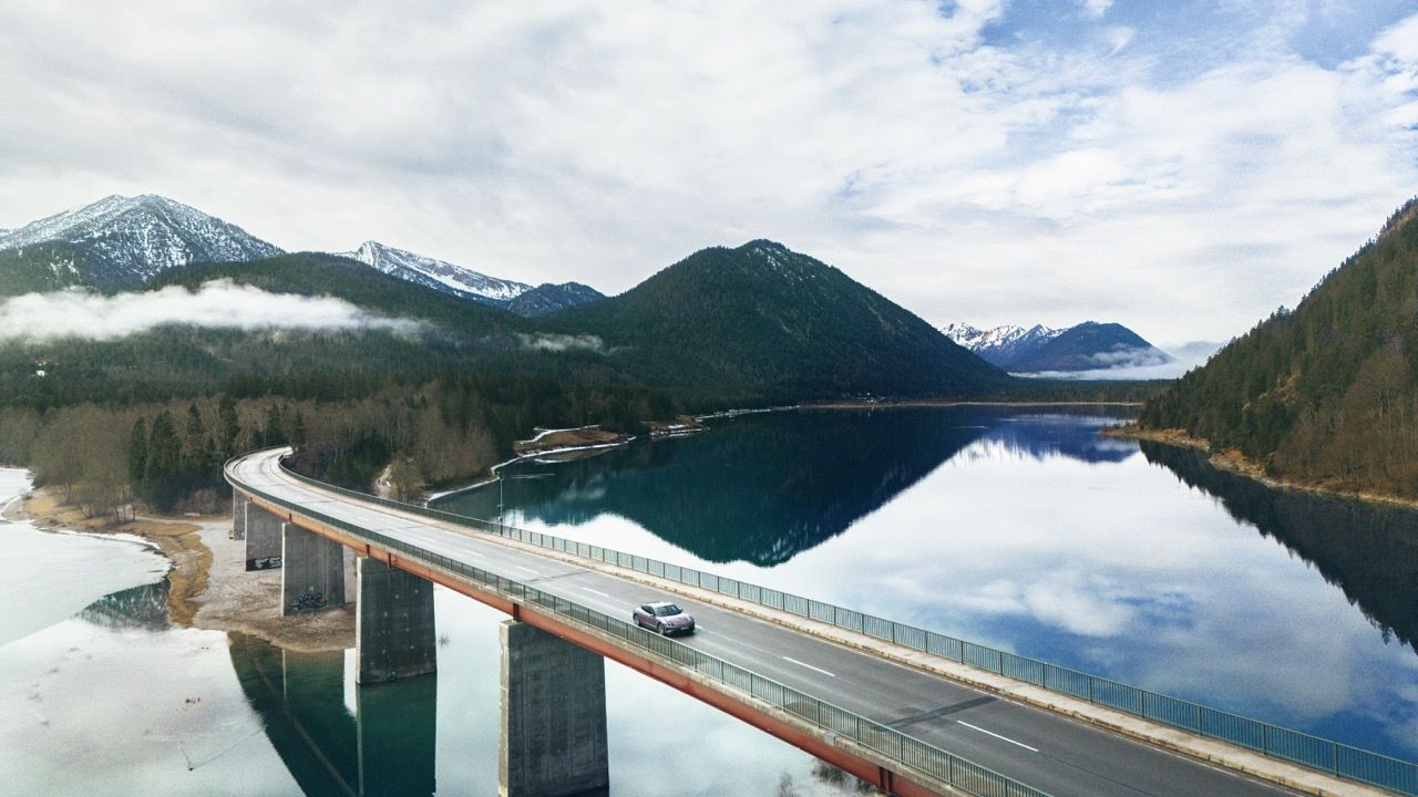 Luftaufnahme eines Porsche Taycan, der eine lange Brücke über einen Alpensee überquert