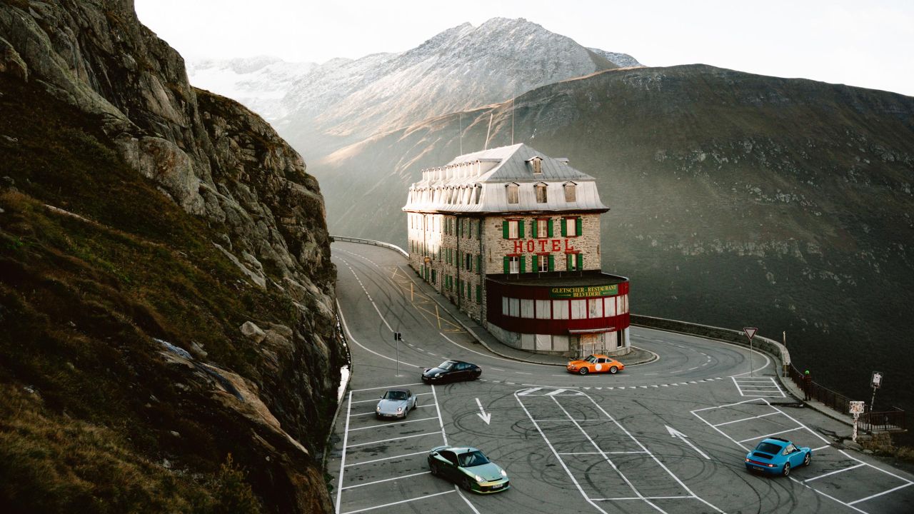 Historic hotel on a sharp bend of a mountain road in the Alpine panorama, with several classic Porsche vehicles parked in its parking lot.