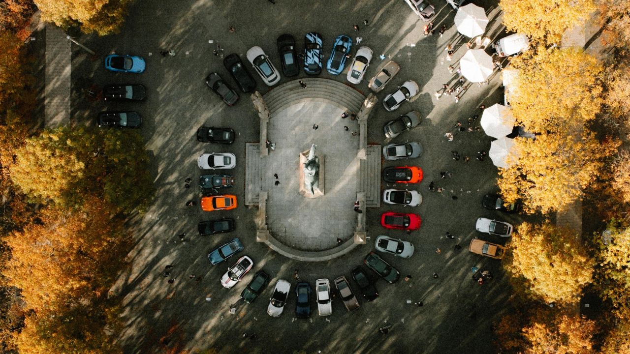 Drone footage shows several Porsche vehicles parked in a circle around a monument in a tree-lined parking lot