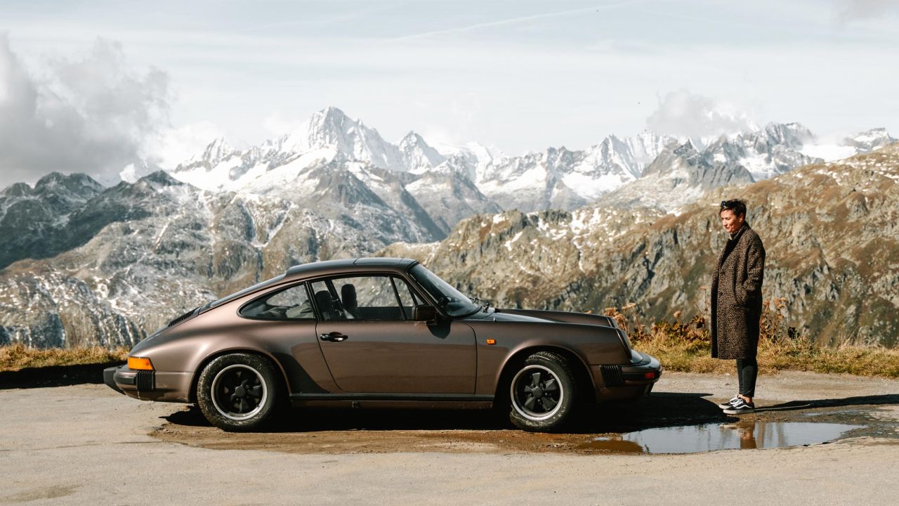 A brown Porsche 911 stands on a mountain road in front of a panorama of snow-covered Alpine peaks, with a woman standing next to the car.
