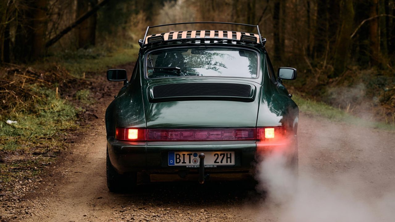 Rear view of a dark green Porsche 911 with roof rack driving on a forest road