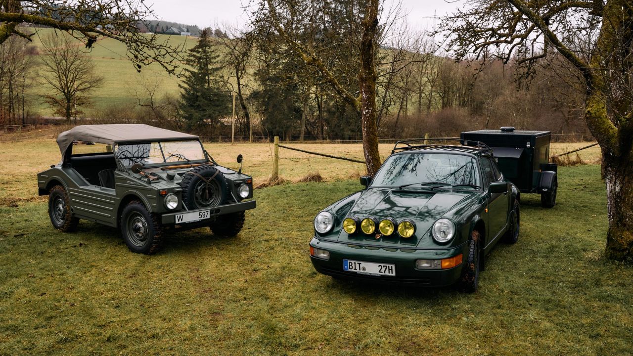 A dark green Porsche 911 with auxiliary headlights and a trailer is parked next to an olive green military off-road vehicle on a meadow.