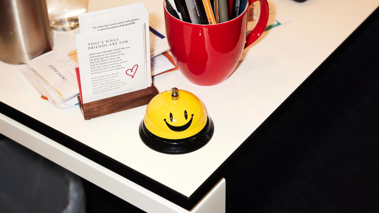 A yellow and black bell with a smiley face is placed on a white desk next to a business card holder and a red pen holder